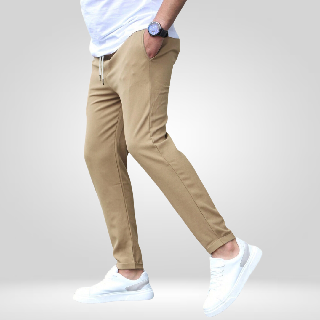 Side profile of tan stretch pants with white sneakers and t-shirt, modeled in a gray studio.