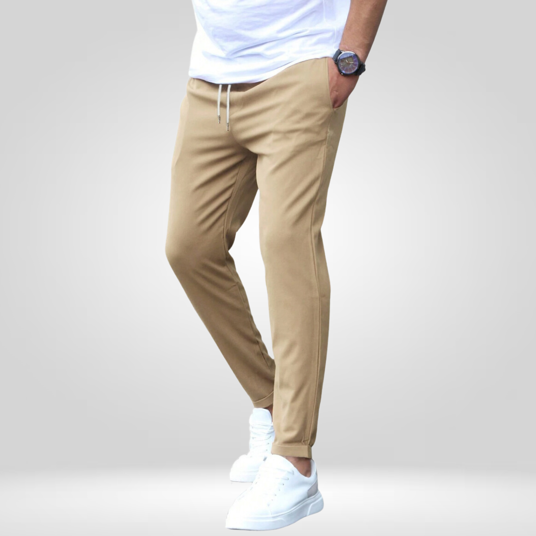 Model wearing tan stretch pants, white t-shirt, and sneakers in a studio with gray backdrop.