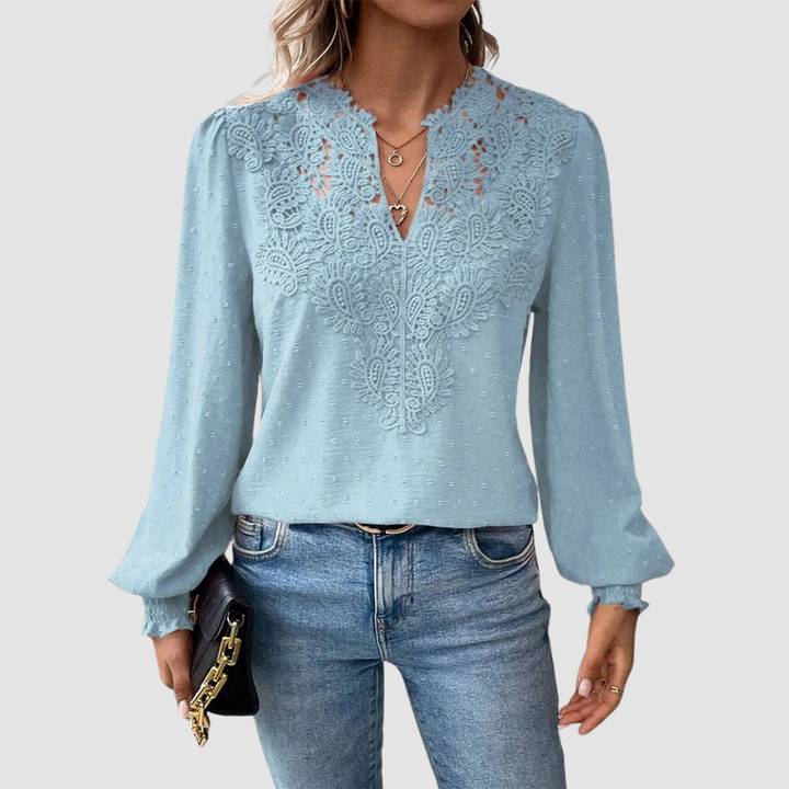 Light blue V-neck lace blouse with intricate design, long sleeves, paired with blue jeans, model holding a black purse, gray background.