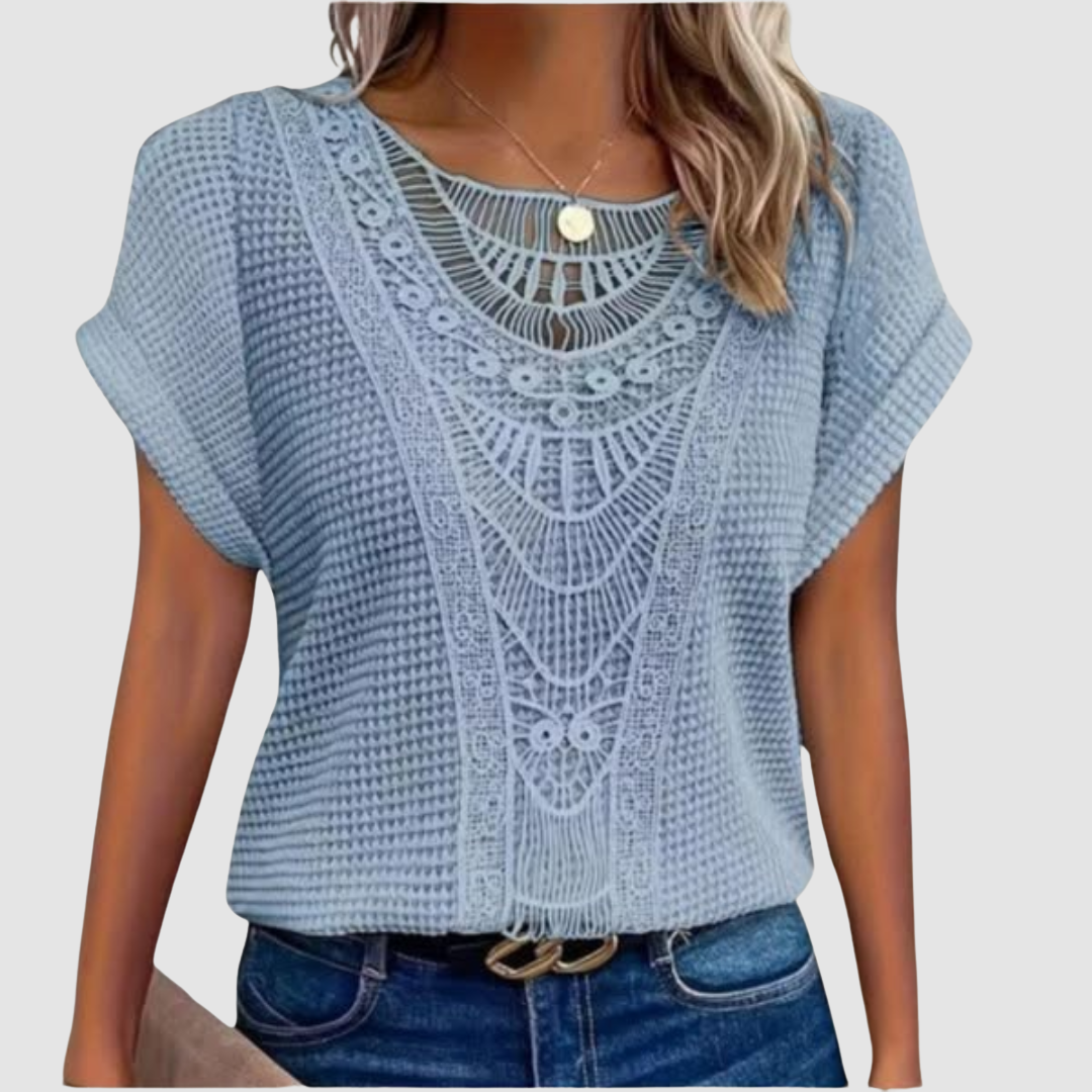 Woman in a light blue lace top with detailed patterns, paired with jeans, studio background.