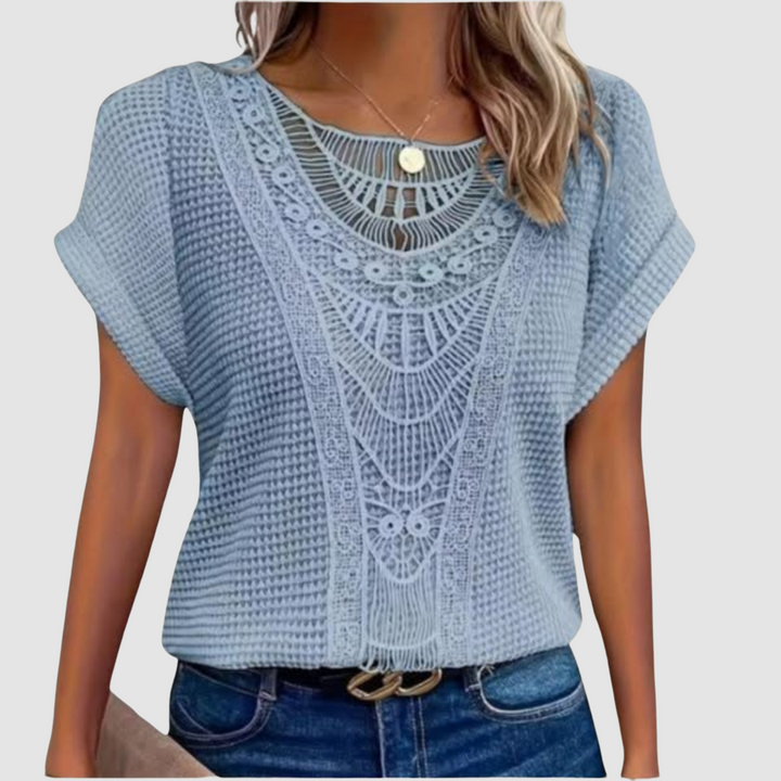 Woman in a light blue lace top with detailed patterns, paired with jeans, studio background.