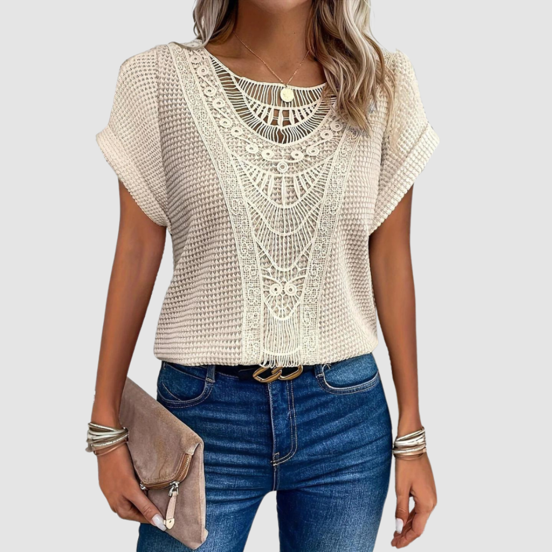 Woman wearing a beige lace top with intricate patterns, paired with jeans and holding a beige clutch, studio background.