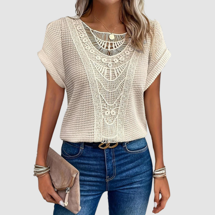 Woman wearing a beige lace top with intricate patterns, paired with jeans and holding a beige clutch, studio background.