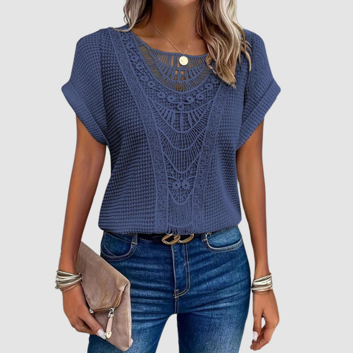 Woman in a navy lace top with detailed patterns, combined with jeans, holding a beige clutch, studio background.