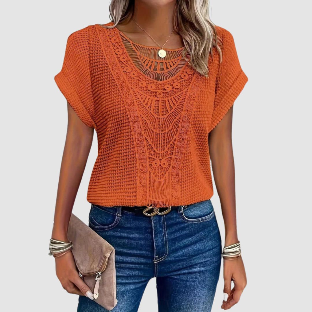 Woman in an orange lace top with intricate patterns, paired with jeans, holding a beige clutch, studio background.