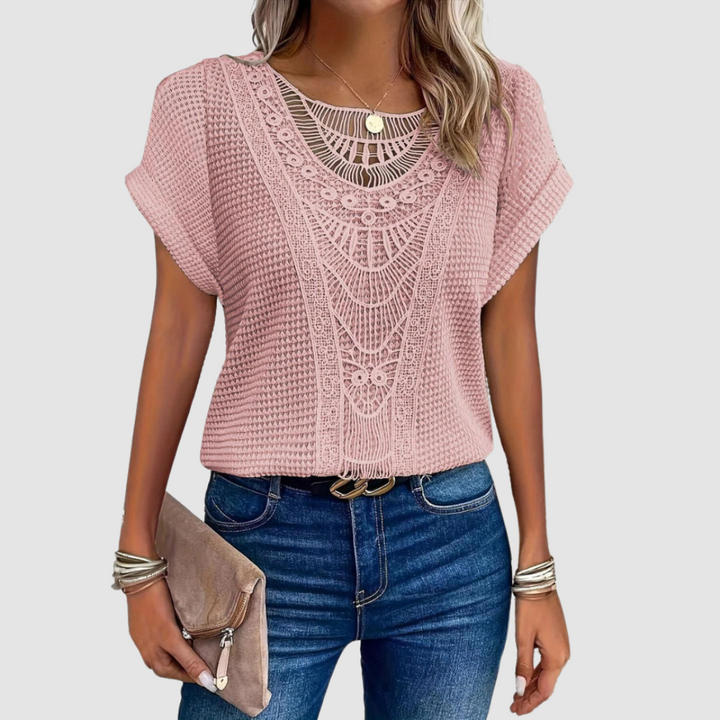 Woman in a pink lace top with delicate patterns, matched with jeans, holding a beige clutch, studio background.