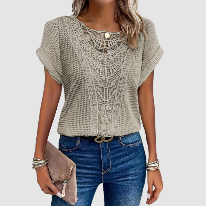 Woman in a taupe lace top with detailed designs, paired with jeans, holding a beige clutch, studio background.