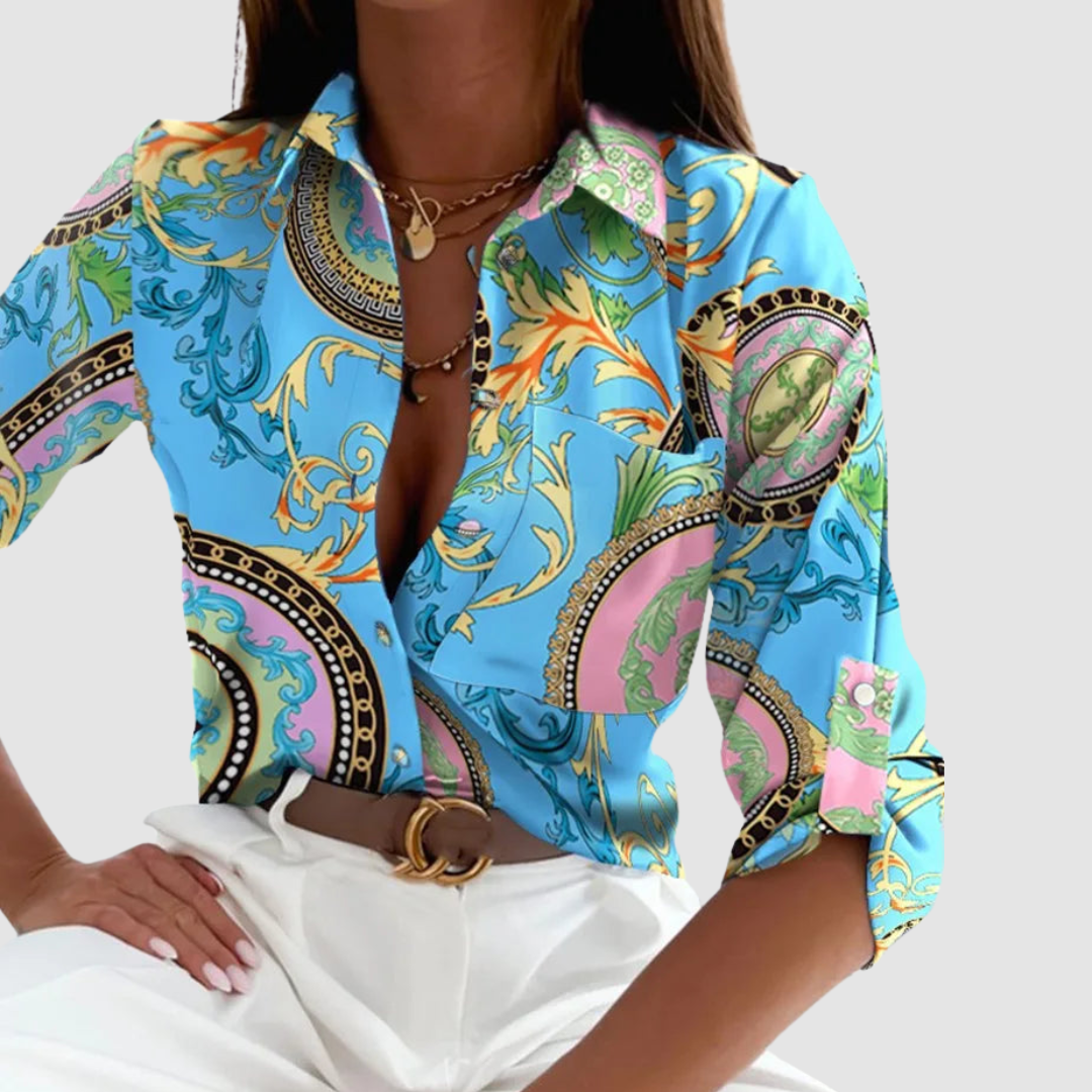 Model in a vibrant blue blouse with ornate patterns, paired with white pants, set against a neutral backdrop.