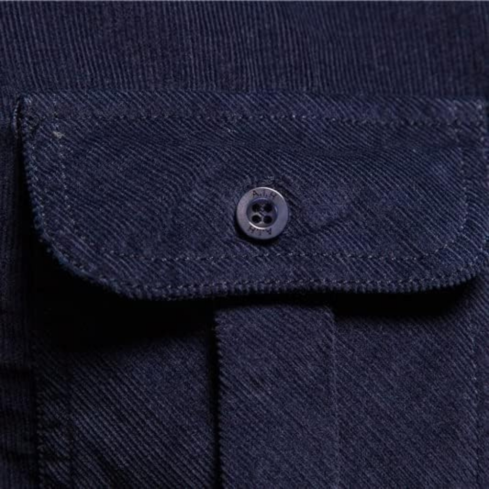 Close-up of the buttoned flap pocket on a dark blue corduroy shirt, showcasing textured fabric and button detail.