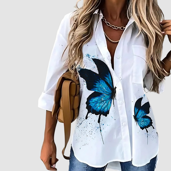 White blouse featuring large blue butterflies, matched with jeans against a plain background.