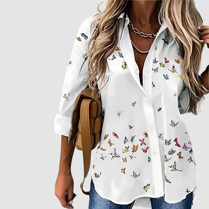 White blouse with small, colorful butterfly accents, paired with jeans against a plain backdrop.