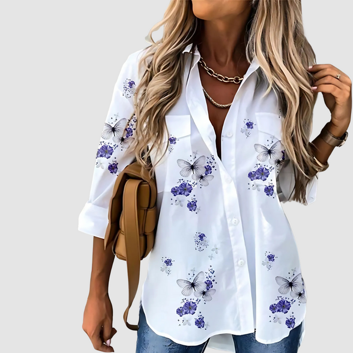 White blouse with blue and purple floral and butterfly design, worn with jeans against a plain background.