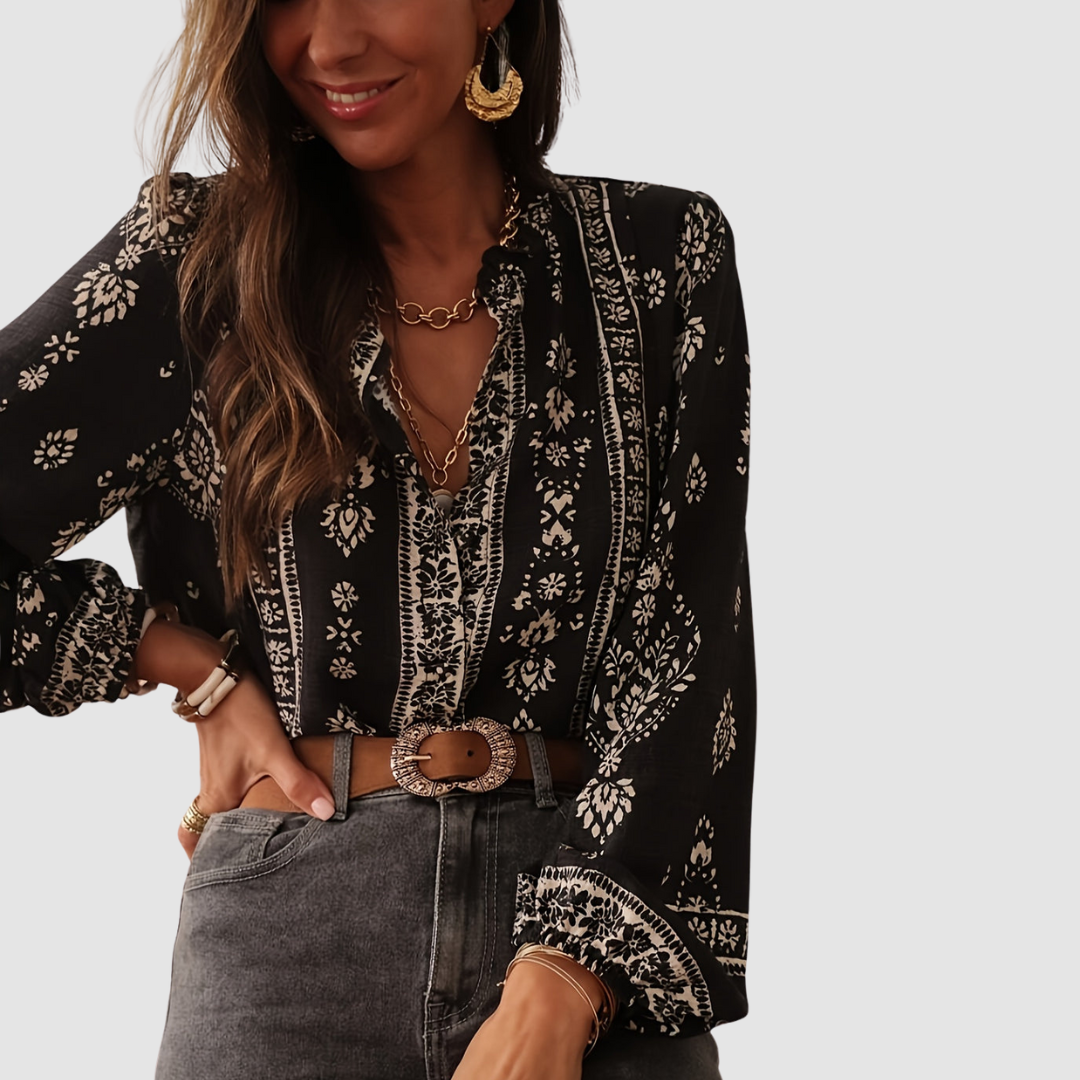 Model in black boho blouse with white floral patterns, gold earrings, and layered necklaces. Studio background.