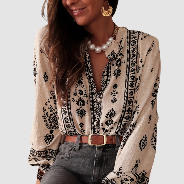 Model wearing a beige boho blouse with black floral patterns, accessorized with chunky jewelry and a brown belt. Studio background.