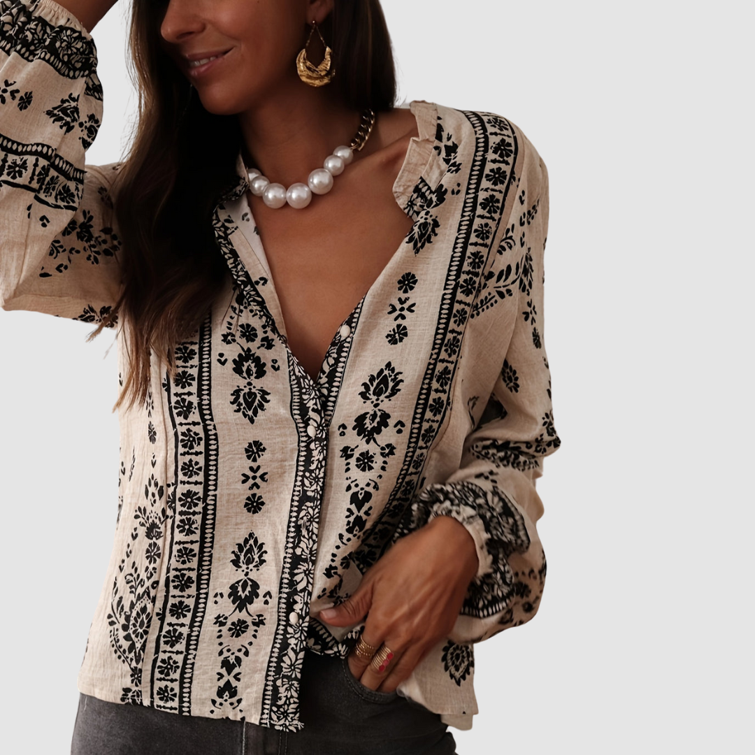 Close-up of a beige boho blouse with black floral designs, styled with pearl necklace and gold jewelry. Studio background.