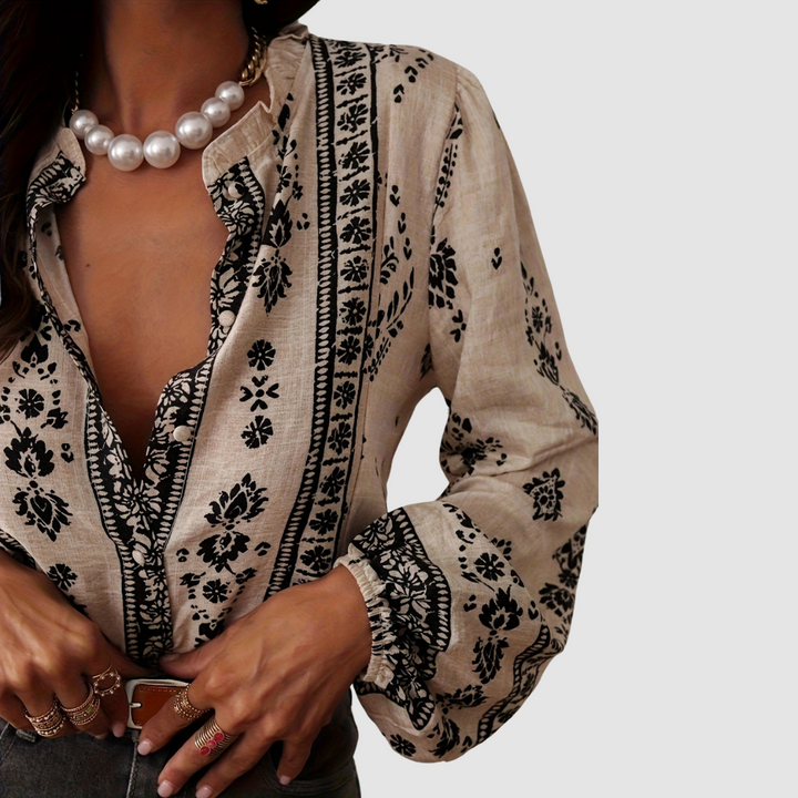 Model in beige boho blouse with intricate black floral print, wearing pearl necklace and multiple rings. Studio setting.