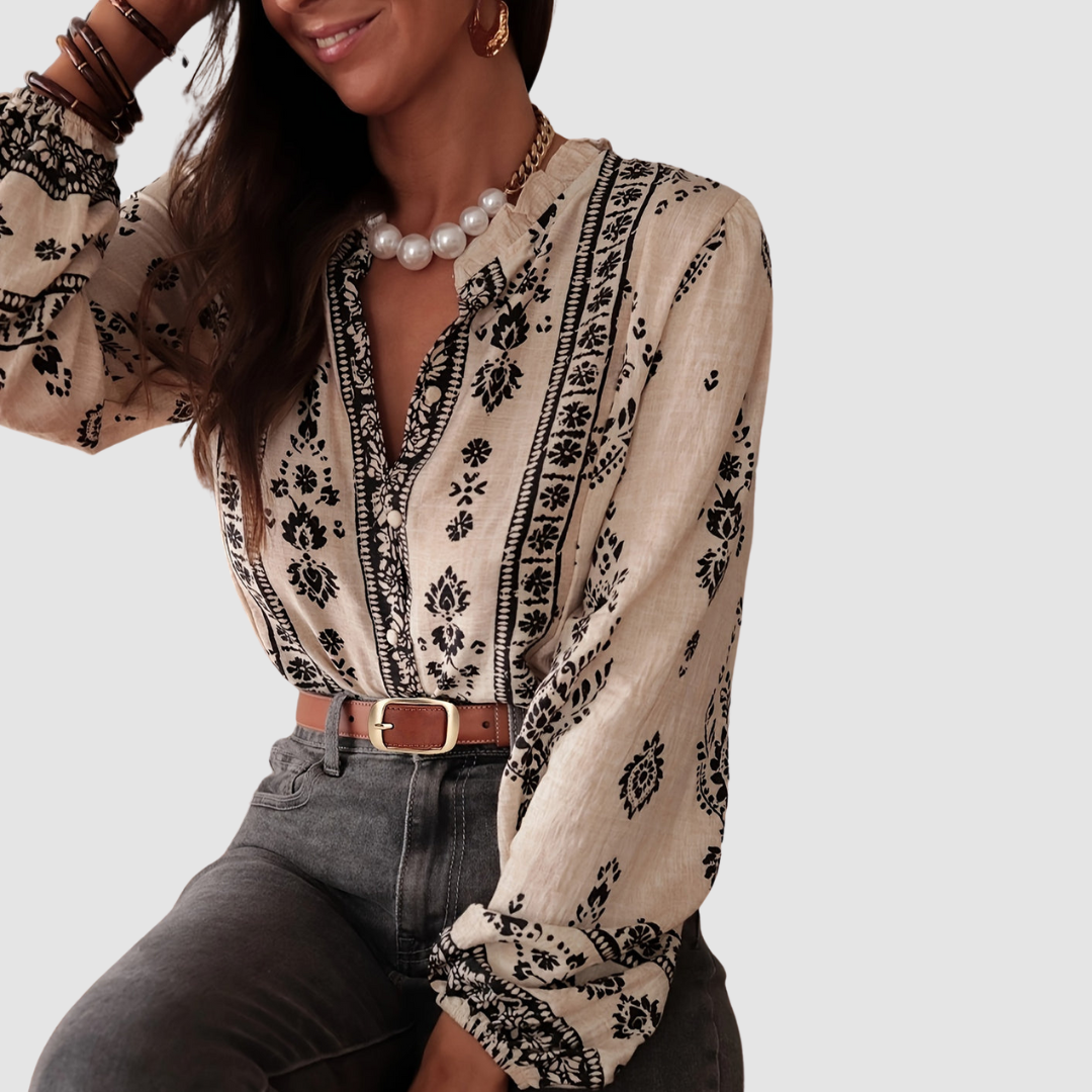 Model sitting in a beige boho blouse with black floral design, accessorized with pearl necklace and brown belt. Studio background.