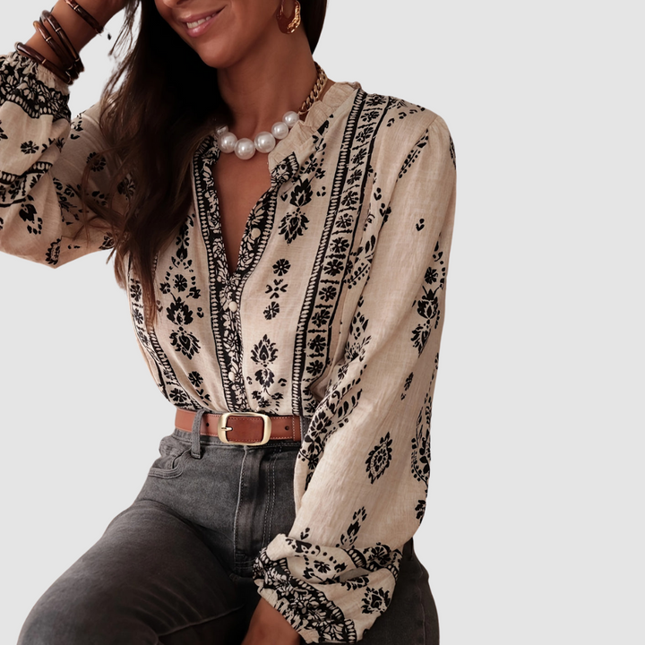 Model sitting in a beige boho blouse with black floral design, accessorized with pearl necklace and brown belt. Studio background.