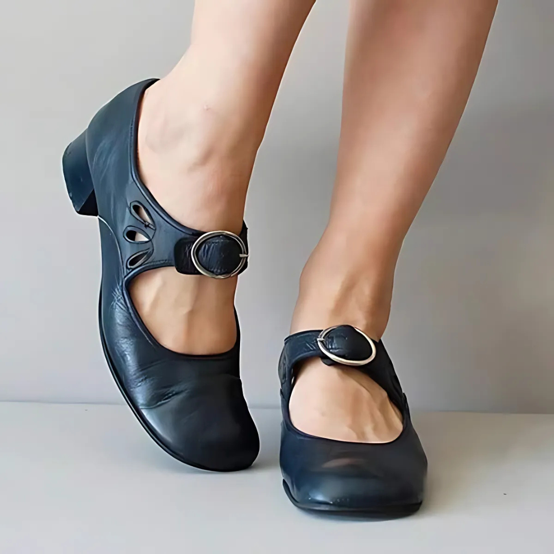 Black leather sandals with buckle detail shown from the front on feet, styled in an elegant setting.