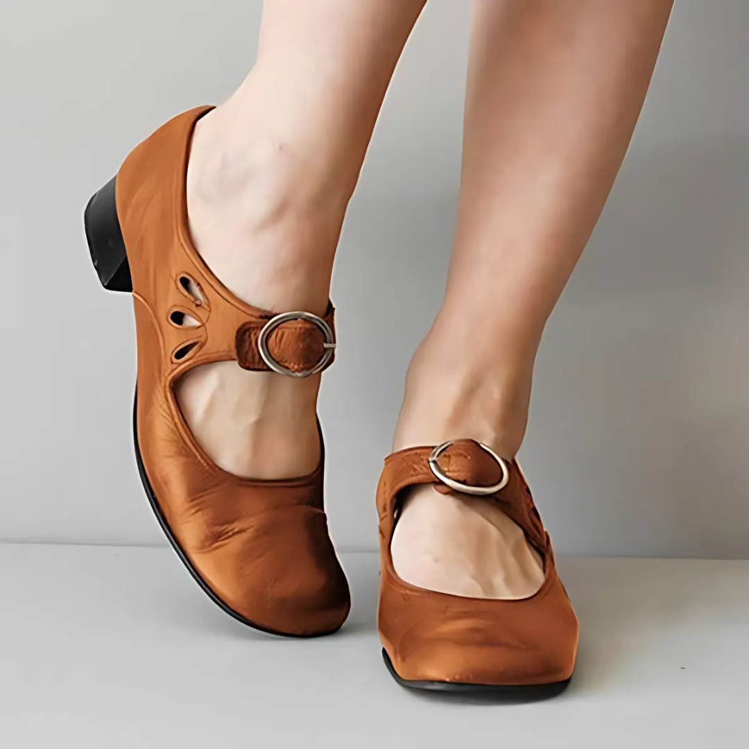 Brown leather sandals with buckle detail shown from the front on feet against a neutral background.