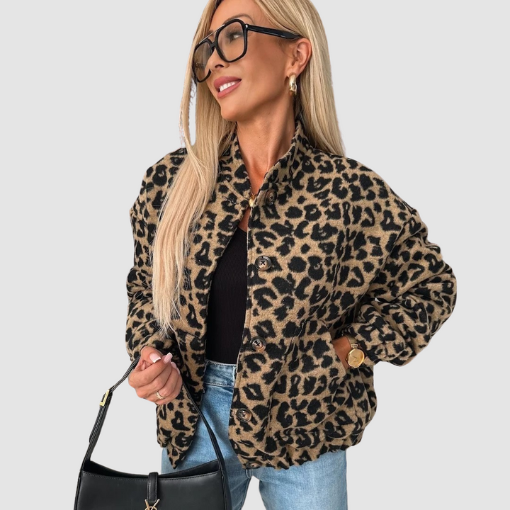 Woman in leopard print jacket and jeans holding a black handbag against a light background.