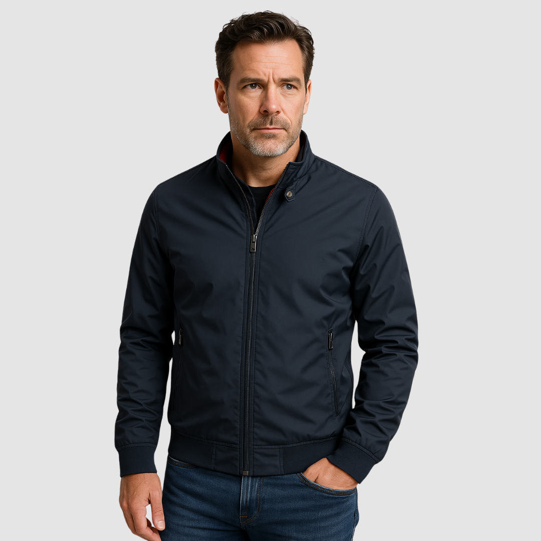 Man wearing a dark blue lightweight bomber jacket, hands in pockets, with a neutral expression against a grey background.