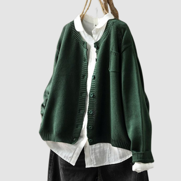 Dark green knit cardigan on hanger over white shirt and dark jeans against light gray background.