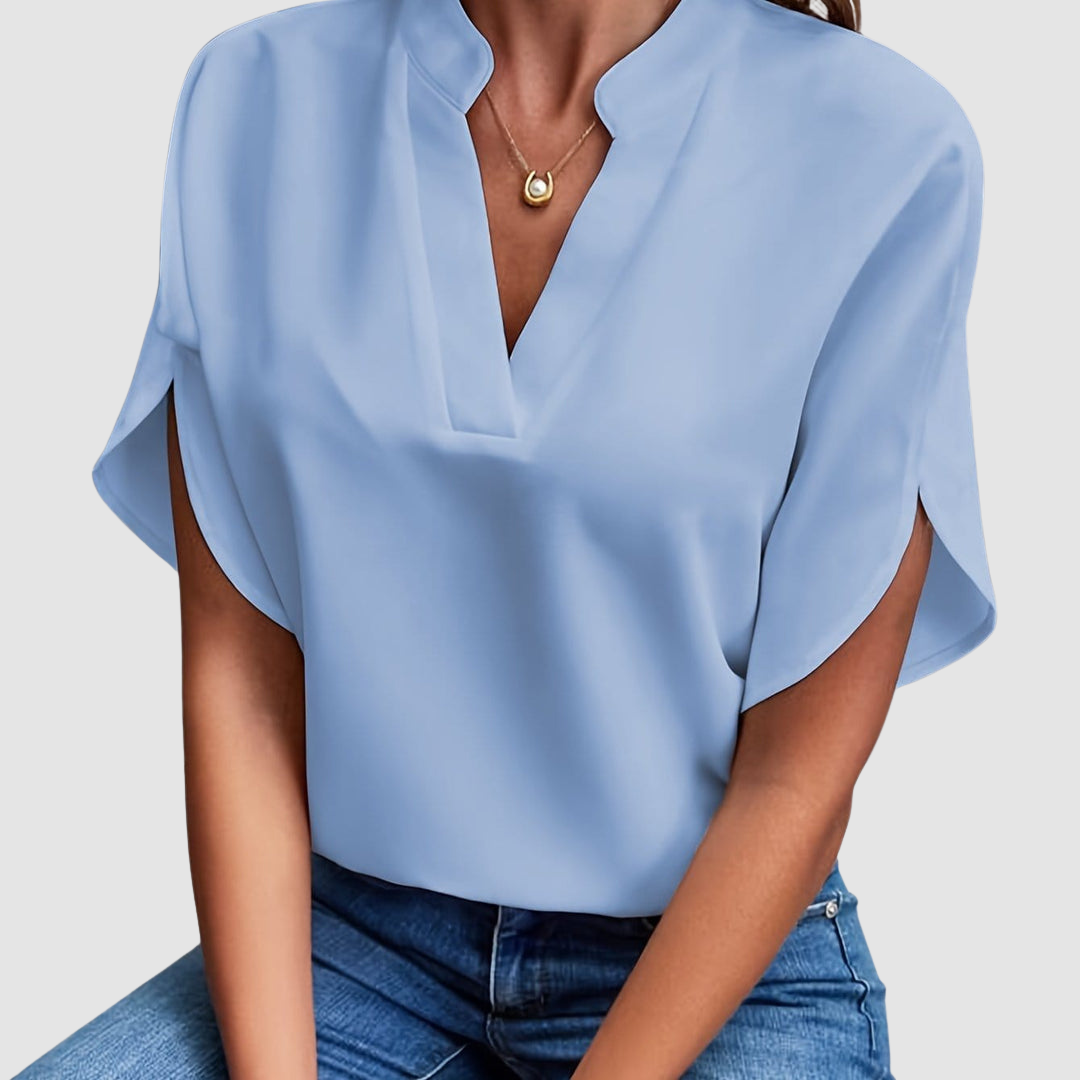 Light blue draped blouse with a sleek V-neck and flowing short sleeves, worn with blue jeans, in a neutral studio setting.