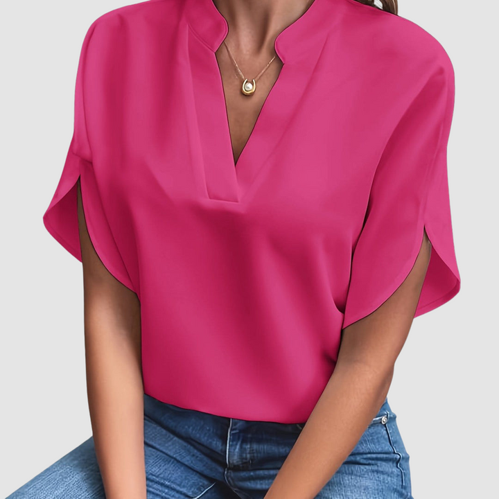 Bright pink draped blouse with a sleek V-neck and flowing short sleeves, worn with blue jeans, in a neutral studio setting.