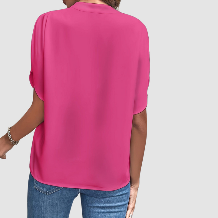 Back view of a bright pink draped blouse, showcasing smooth, flowing fabric and easy fit, paired with blue jeans, studio setting.