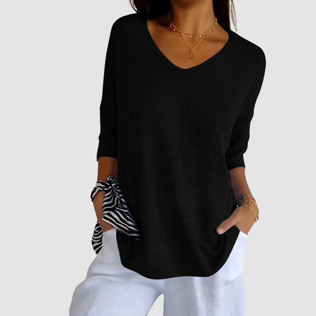 Black knit V-neck top with short sleeves, worn with white pants. Model with hands in pockets against plain background.