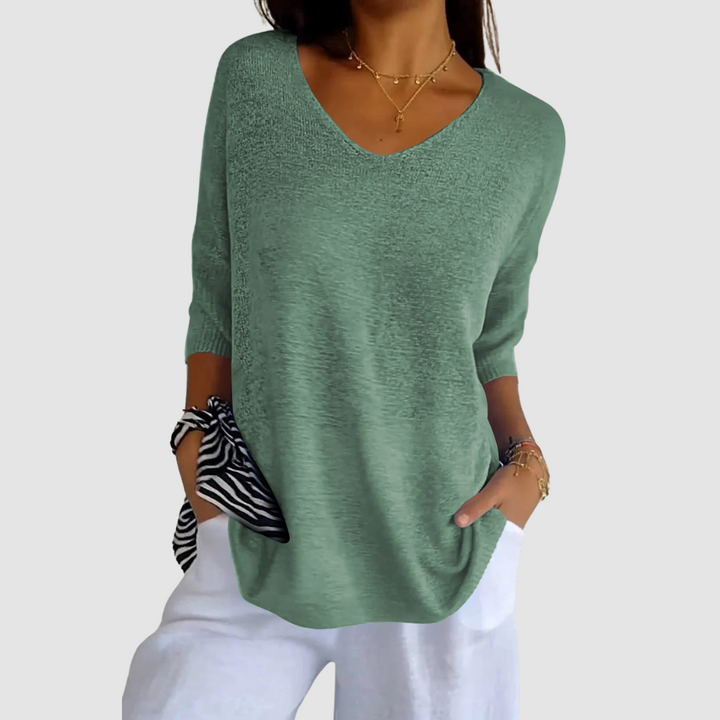 Light green knit V-neck top with short sleeves, matched with white pants. Model posing hands in pockets, plain background.