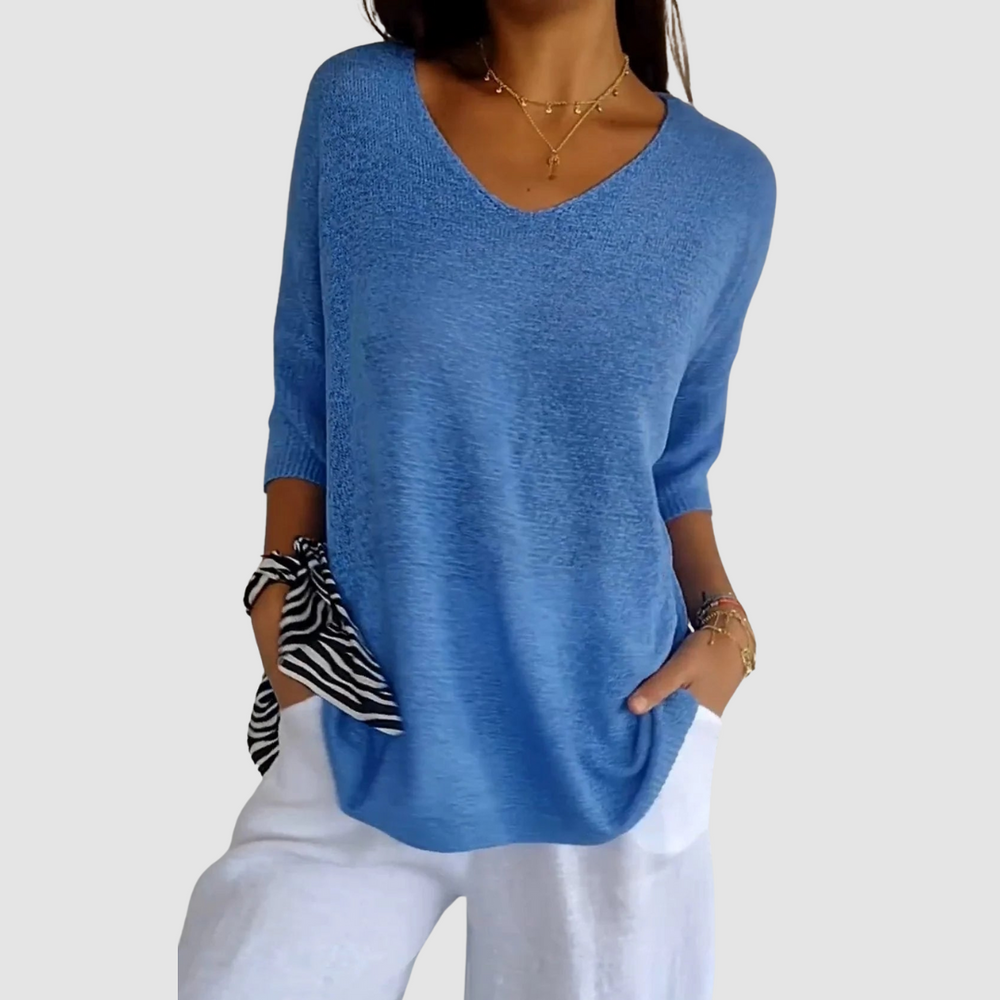 Blue knit V-neck top with short sleeves, paired with white pants. Model posing hands in pockets, plain backdrop.