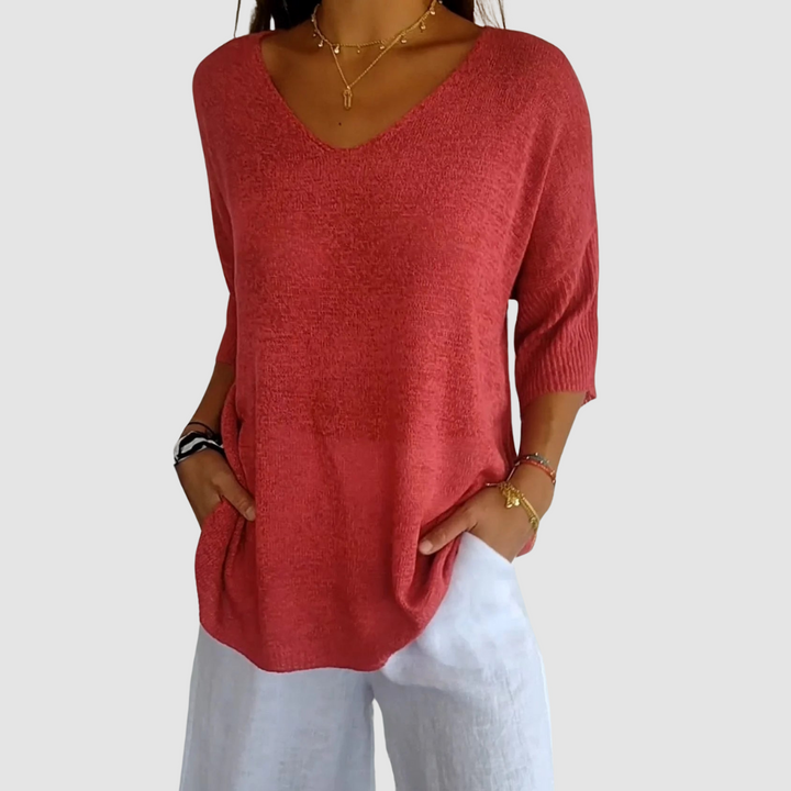 Red knit V-neck top with short sleeves, matched with white pants. Model with hands in pockets against plain background.