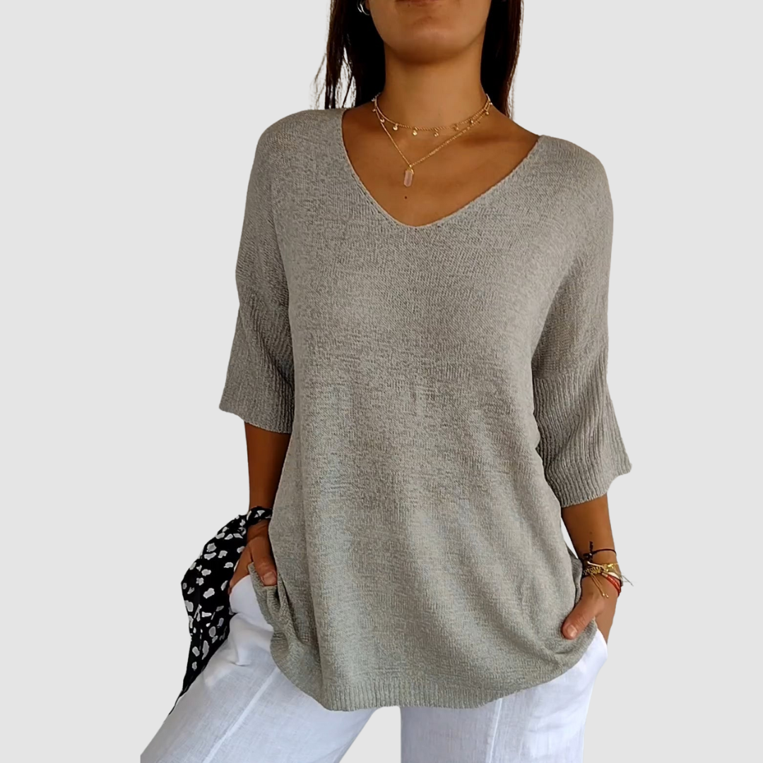 Gray knit V-neck top with short sleeves, worn over white pants. Model has hands in pockets, plain background.