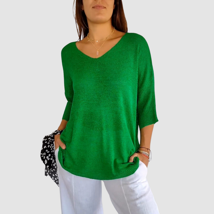 Green knit V-neck top with short sleeves, worn with white pants. Model has hands in pockets, plain background.