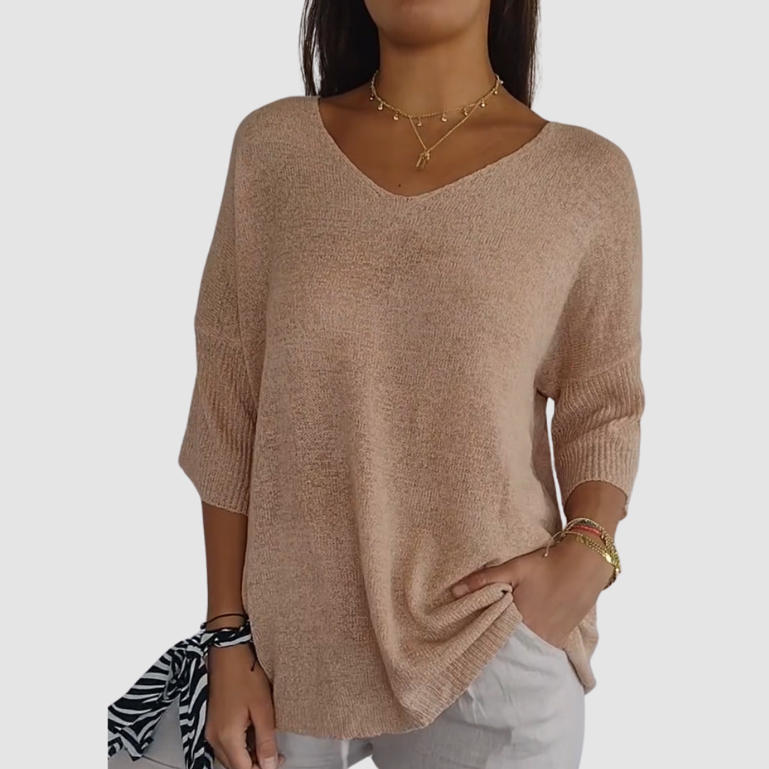 Beige knit V-neck top with short sleeves, worn with white pants. Model has hands in pockets, plain background.