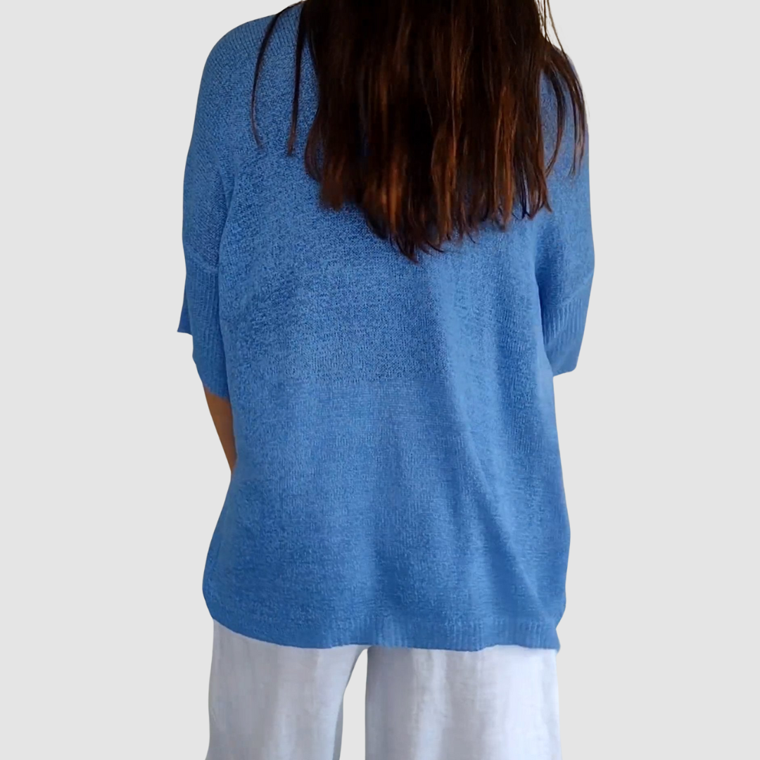 Back view of blue knit V-neck top with short sleeves worn over white pants, plain background.