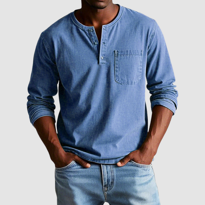 Man wearing a blue vintage Henley shirt, hands in pockets, in a studio setting against a light background.