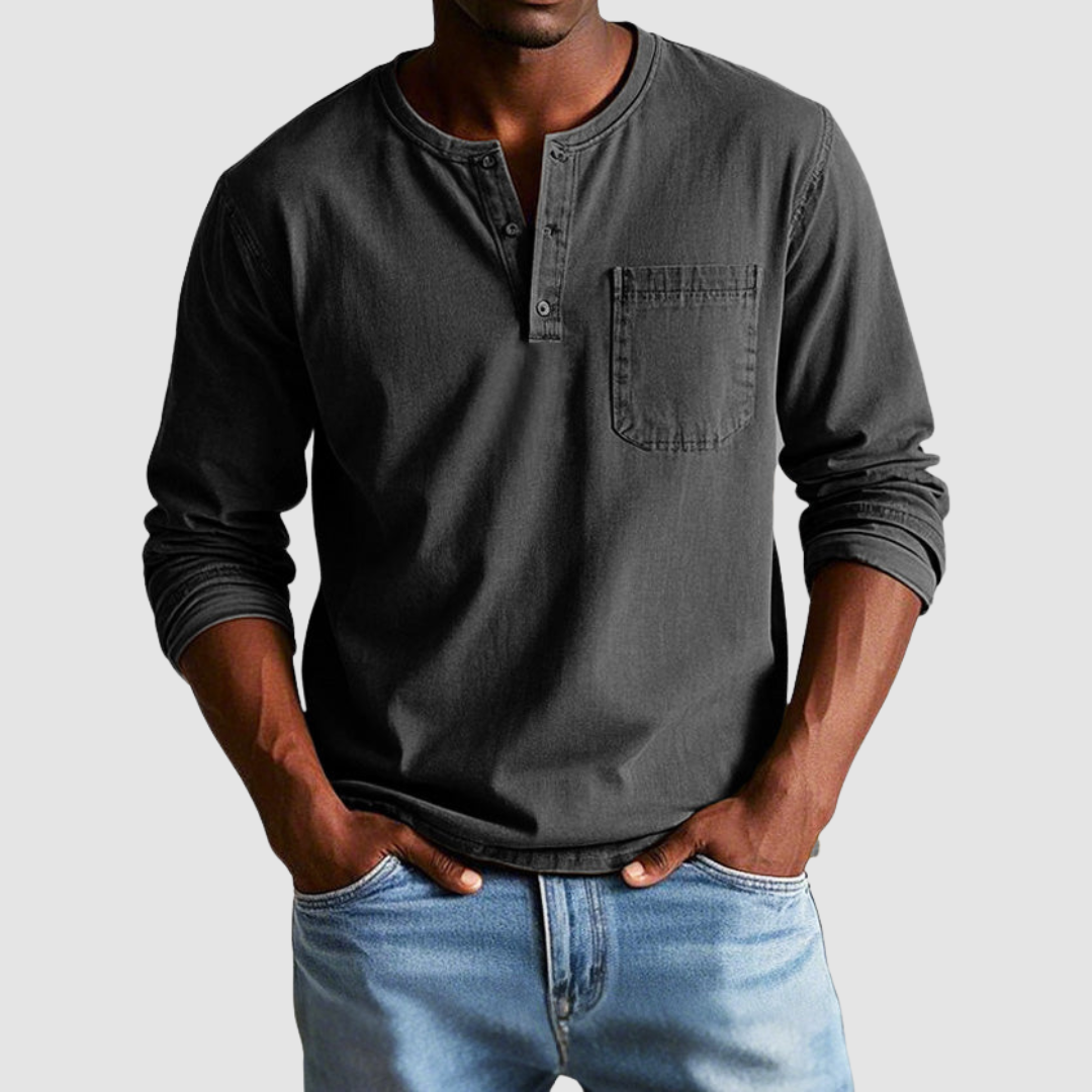 Man wearing a dark gray vintage Henley shirt, hands in pockets, in a studio setting against a light background.