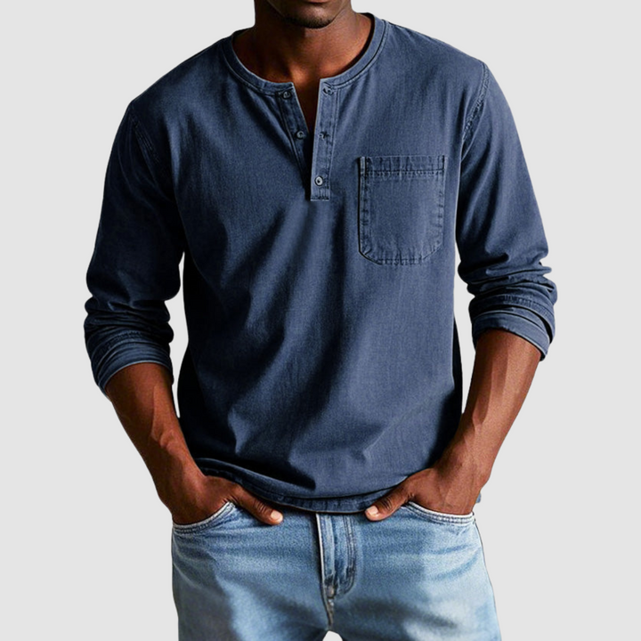 Man wearing a dark blue vintage Henley shirt, hands in pockets, in a studio setting against a light background.