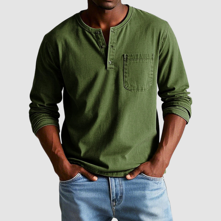 Man wearing a dark green vintage Henley shirt, hands in pockets, in a studio setting against a light background.