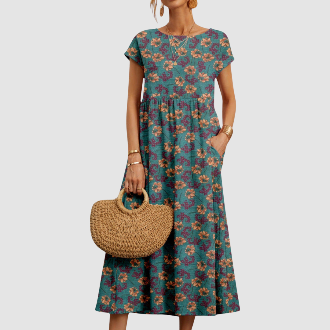 Teal floral midi dress with orange and purple flowers, short sleeves, complemented by a straw handbag, on a light gray background.