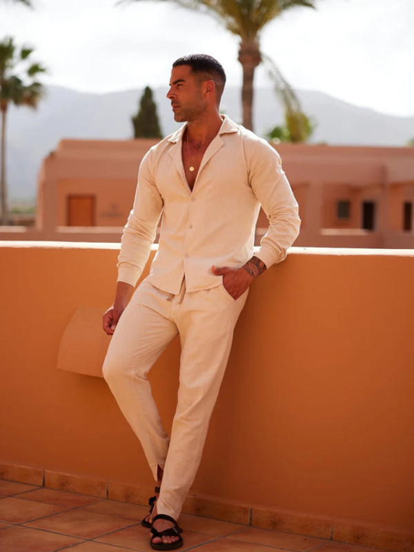 Model in cream-colored summer set, leaning against a warm-toned wall with a scenic backdrop of mountains and palm trees.