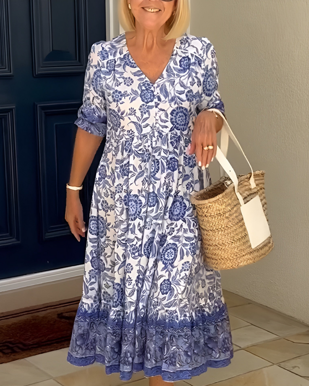 Woman wearing a blue and white floral V-neck dress, holding a straw bag in front of a navy door.