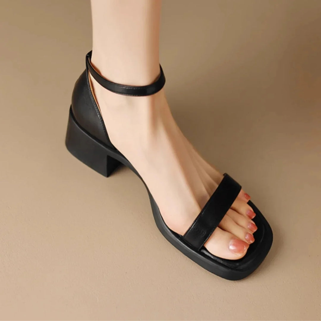 Black leather sandal with ankle strap and block heel on foot, placed on neutral studio floor.