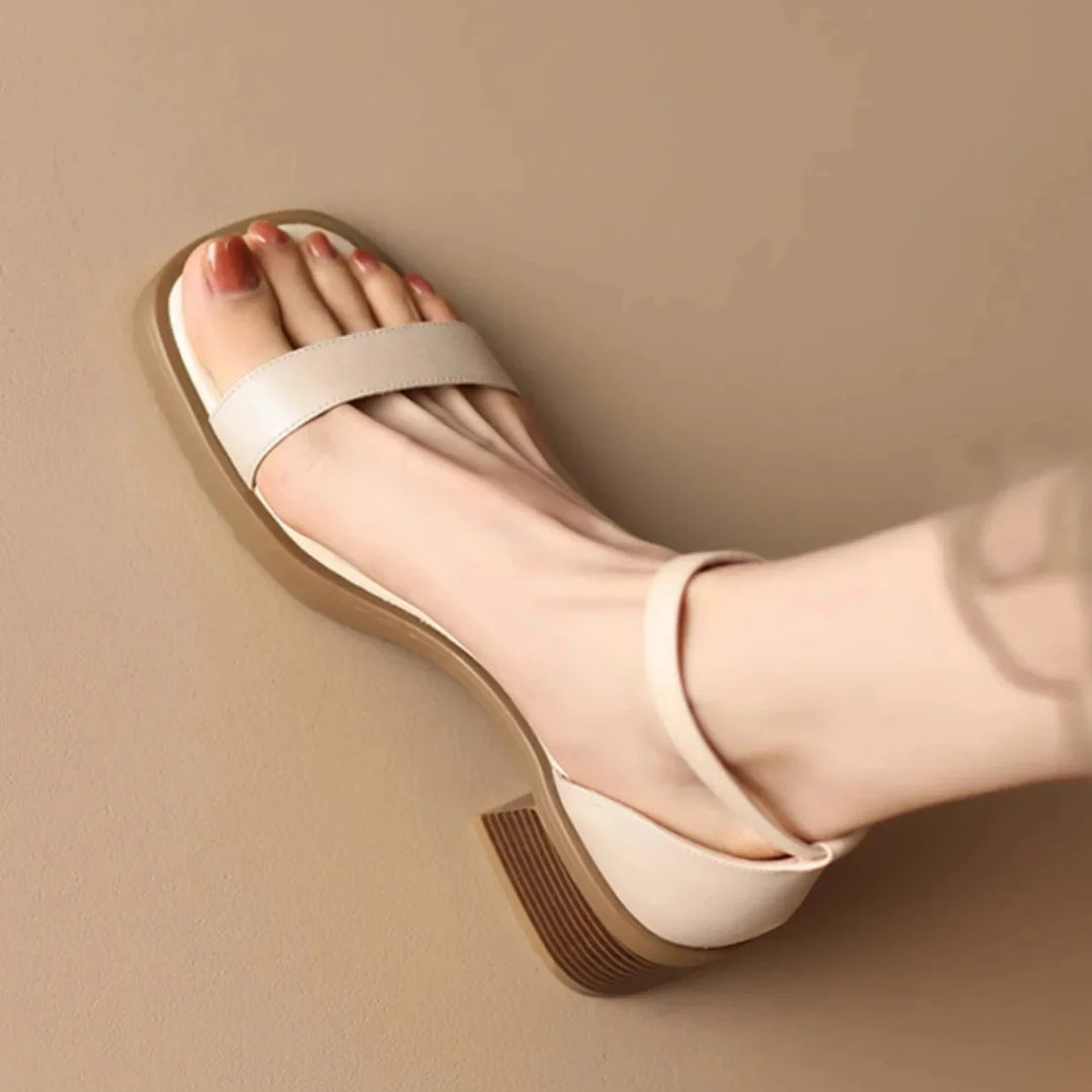 Light cream leather sandal with block heel on foot, leaning against neutral studio wall.