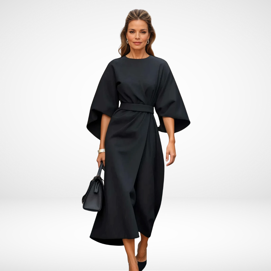 Model in elegant black midi dress with wide sleeves and waist tie, holding a black handbag in a studio setting.