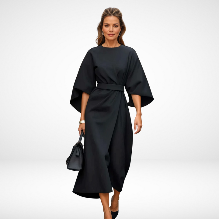 Model in elegant black midi dress with wide sleeves and waist tie, holding a black handbag in a studio setting.