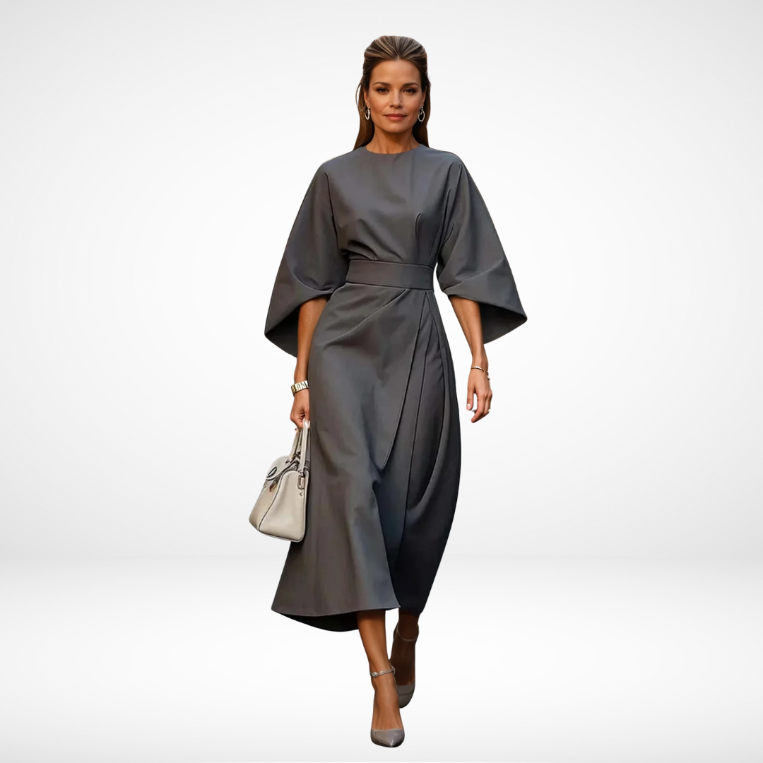 Model in stylish grey midi dress with wide sleeves and waist tie, holding a light handbag in a studio environment.